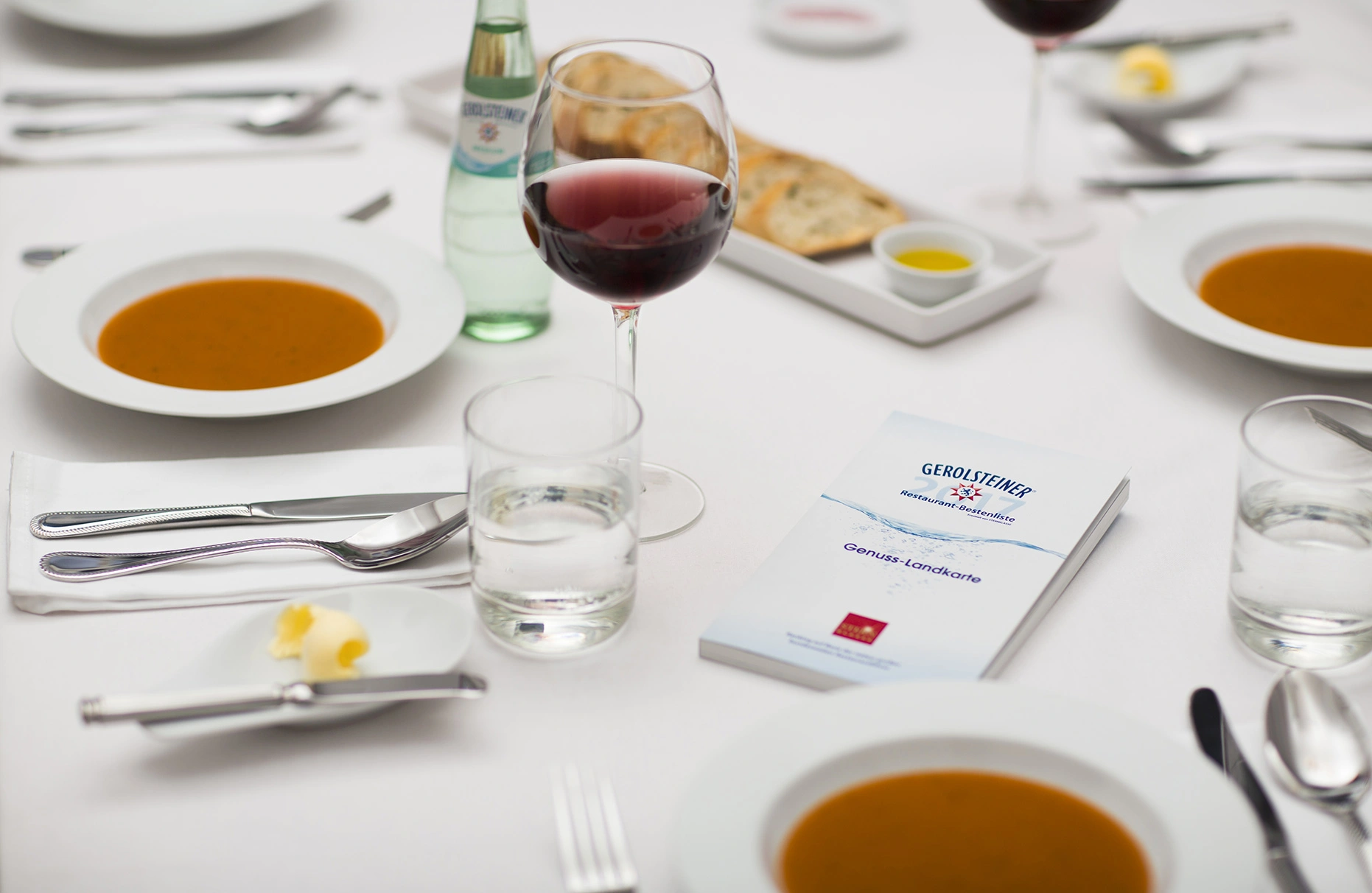 Auf einem weissen gedeckten Tisch mit gefüllten Suppenteller und gefüllten Rotwein Gläsern liegt ein Buch namens Gerolsteiner Genuss Landkarte. Produziert für die PR-Agentur Schröder und Schömbs PR aus Berlin.