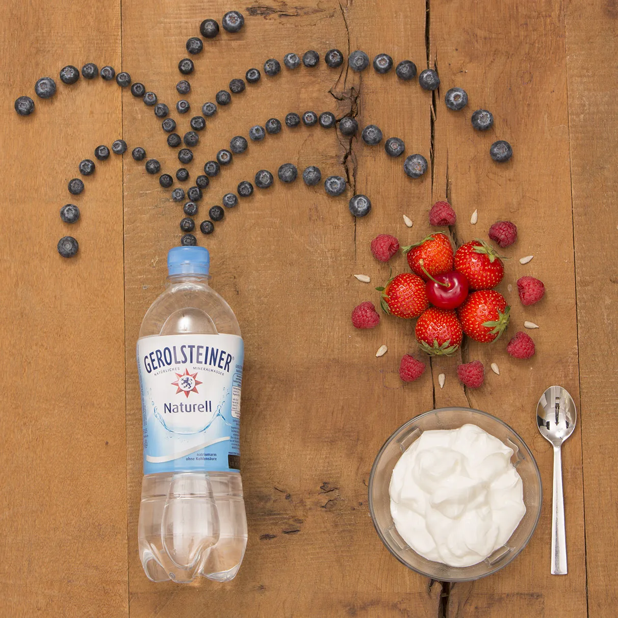 Auf einem natürlichen Holzuntergrund liegt links eine Gerolsteiner PET Flasche aus der Blaubeeren sprudeln. Die Blaubeeren sind dabei jedoch nur so hingelegt, dass es nach Sprudeln ausieht. Rechts daneben steht eine Joghut Schale aus Glas mit Löffel und darüber etwas Obbst, welches Sternförmig gelegt wurde. Das ganze ist von oben im flat lay Stil fotografiert . Produziert für die PR-Agentur Schröder und Schömbs PR aus Berlin.