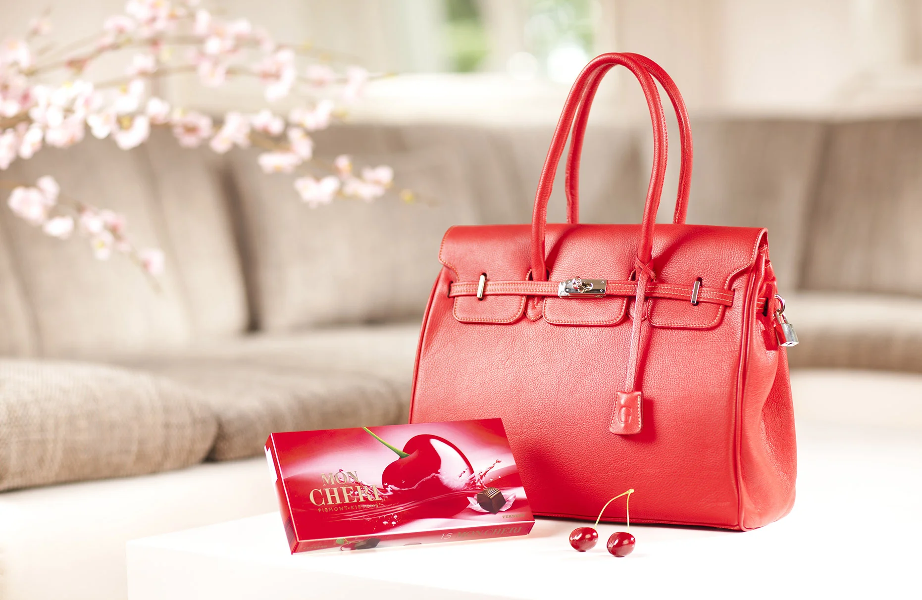 Auf einem weissen Couchtisch steht eine Schachtel Mon Cheri und eine knallrote Damenhandtasche. Im Hintergrund zu sehen eine beige farbene Couch und eine hereinragender blühender Kirschbaumzweig. Produziert für die PR-Agentur Schröder und Schömbs PR aus Berlin.