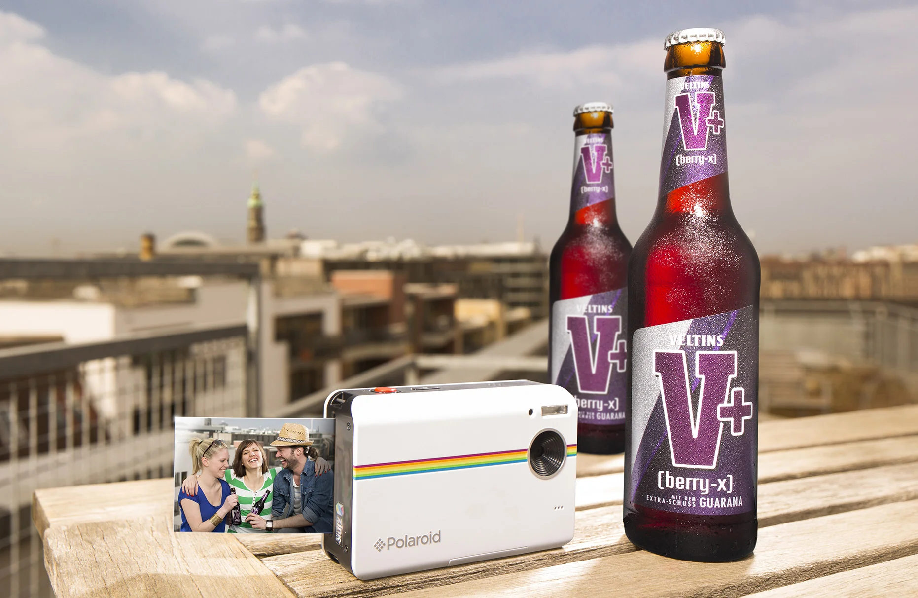 Über den Dächern einer Stadt stehen auf einem Balkon- Tisch zwei eiskalte Veltins V+ Flaschen mit Wasserperlen und links daneben eine kleine modernere Polaroid Kamera. Produziert für die PR-Agentur Schröder und Schömbs PR aus Berlin.